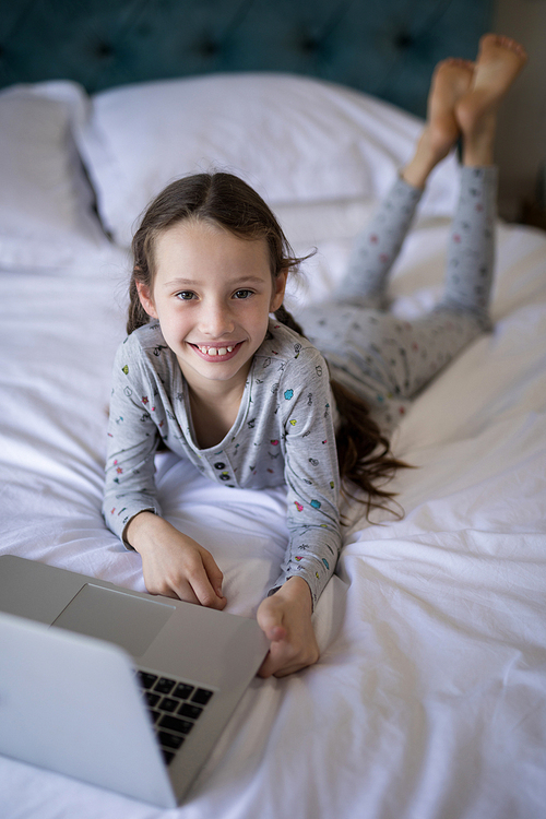 Portrait of smiling girl lying on bed with laptop in bedroom