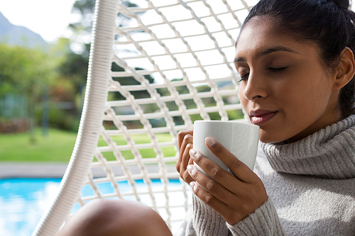 Woman holding coffee cup while relaxing in swing chair at poolside
