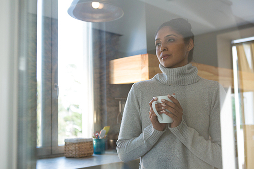 Thoughtful woman with coffee cup seen through glass in kitchen at home