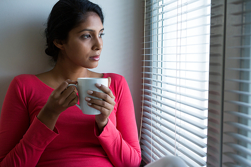 Thoughtful young woman with coffee cup by window at home