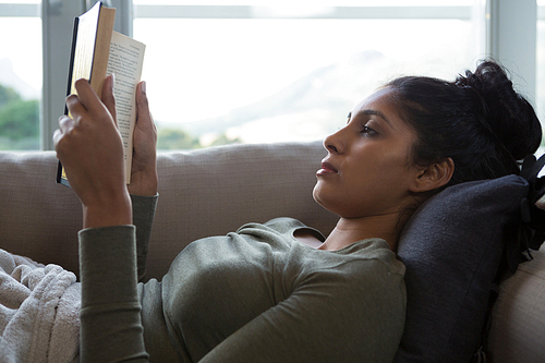 Young woman reading book while relaxing on sofa at home