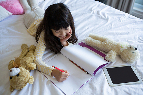 Relaxed girl with toys writing in book on bed at home