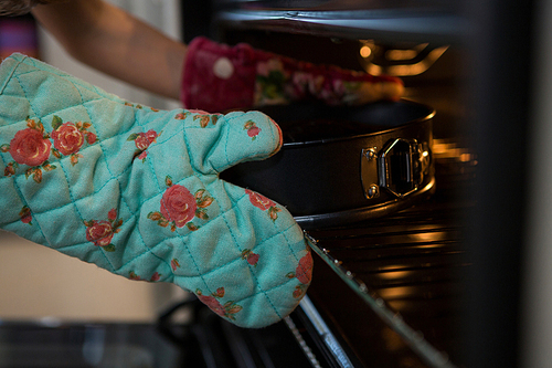 Cropped hands of girl wearing glove keeping container with cake in oven at kitchen