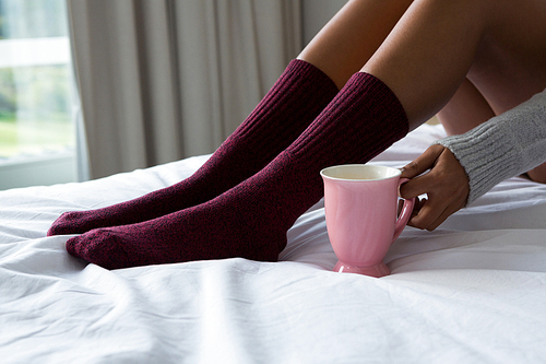 Low section of woman in sock holding coffee cup on bed at home