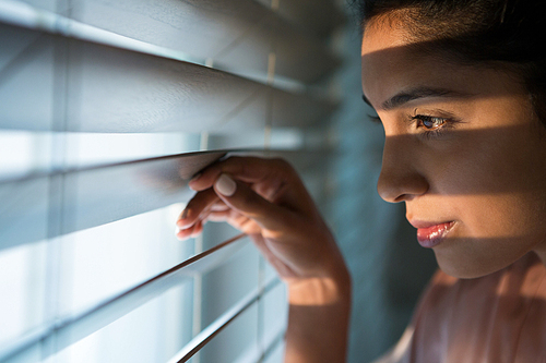 Close-up of thoughtful young woman looking through window blinds