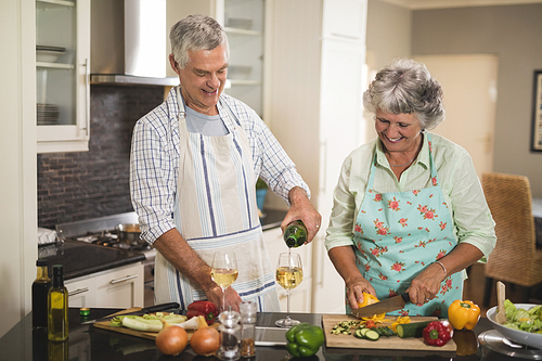 Smiling senior man pouring wine in glass while woman cutting vegetables at kitchen counter