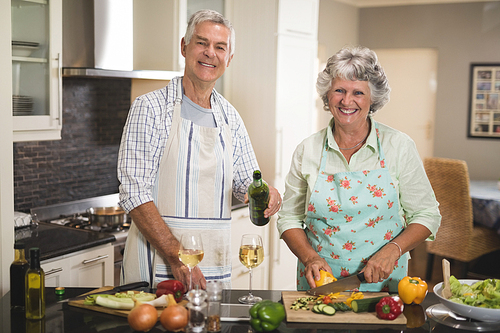 Portrait of smiling senior man holding wine bottle by woman cutting vegetables at kitchen counter