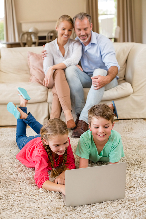 Sibling lying on rug and using laptop in living room while parents sitting in background at home