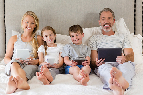 Portrait of smiling parents and kids using mobile phone and digital tablet on bed in bedroom