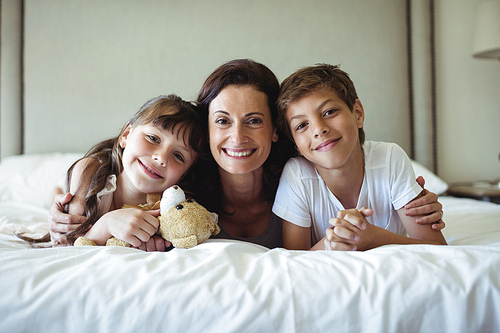 Portrait of smiling mother and kids lying on bed with teddy bear in bedroom