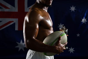 Mid section of shirtless man with trophy and rugby ball standing against australian flag