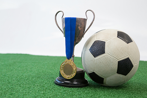 Close-up of football, trophy and medal on artificial grass against white background