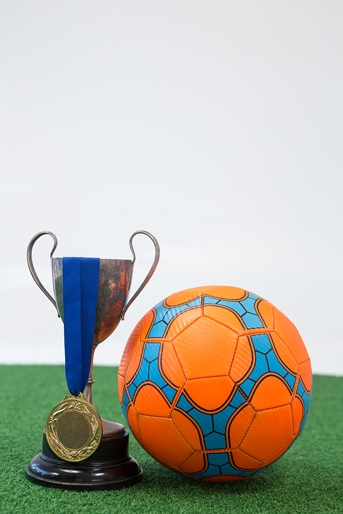 Close-up of football, trophy and medal on artificial grass against white background