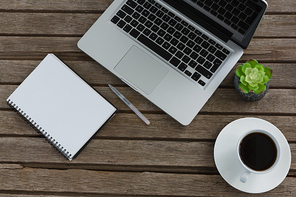 Overhead of laptop, notepad, pen, pot plant, and coffee on wooden plank