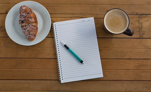 Overhead of croissant, coffee, pen, notepad on wooden plank