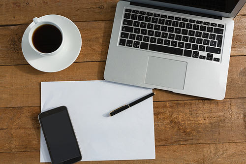 Overhead of laptop, mobile phone, black coffee, pen and paper on wooden plank