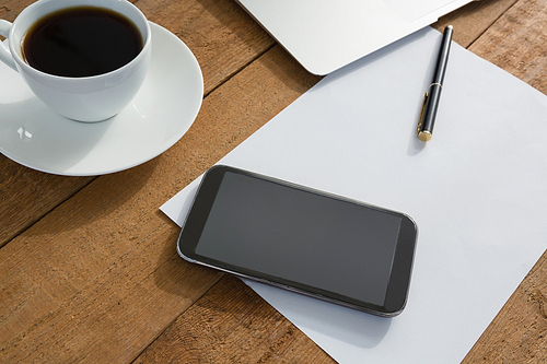 Close-up of laptop, mobile phone, black coffee, pen and paper on wooden plank