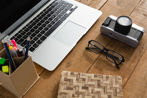 Close-up of laptop, camera, spectacles, notepad and pencil holder on wooden plank