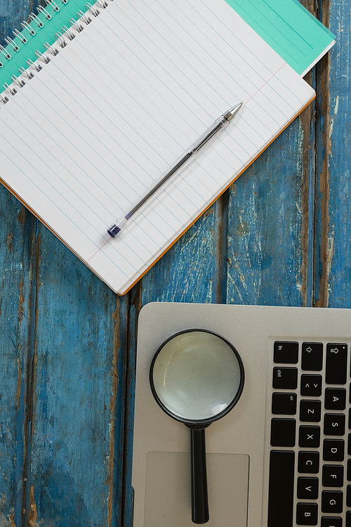 Overhead of blank book, pen, magnifying glass and laptop on wooden plank