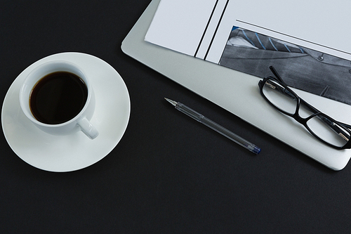 Overhead of black coffee, spectacles, pen, laptop and newspaper on black background