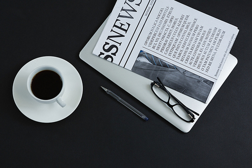 overhead of  coffee, spectacles, pen, laptop and newspaper on black background