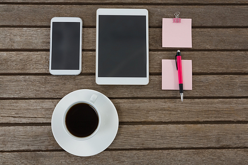 Overhead of black coffee, mobile phone, digital tablet, pen and sticky note on wooden plank