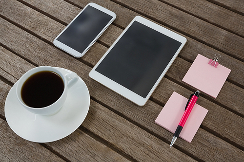 Overhead of black coffee, mobile phone, digital tablet, pen and sticky note on wooden plank