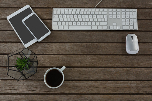 Overhead of digital tablet, mobile phone, mouse, pot plant, keyboard and black coffee on wooden plank