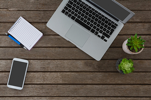 Overhead of laptop, pot plant, notepad, pen and mobile phone on wooden plank