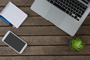 Overhead of laptop, pot plant, notepad, pen and mobile phone on wooden plank