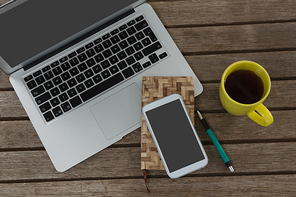 Overhead of laptop, mobile phone, dairy, pen and coffee on wooden plank