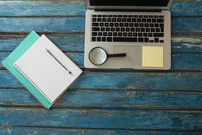 Overhead of blank book, pen, magnifying glass and laptop on wooden plank