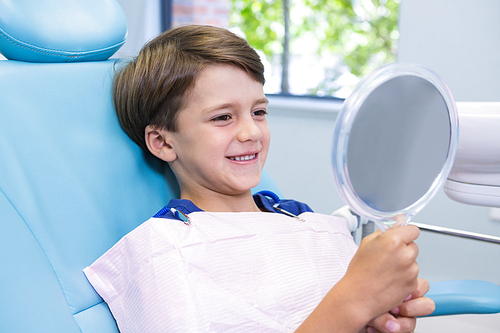 Cute boy looking at mirror while sitting on chair at dental clinic