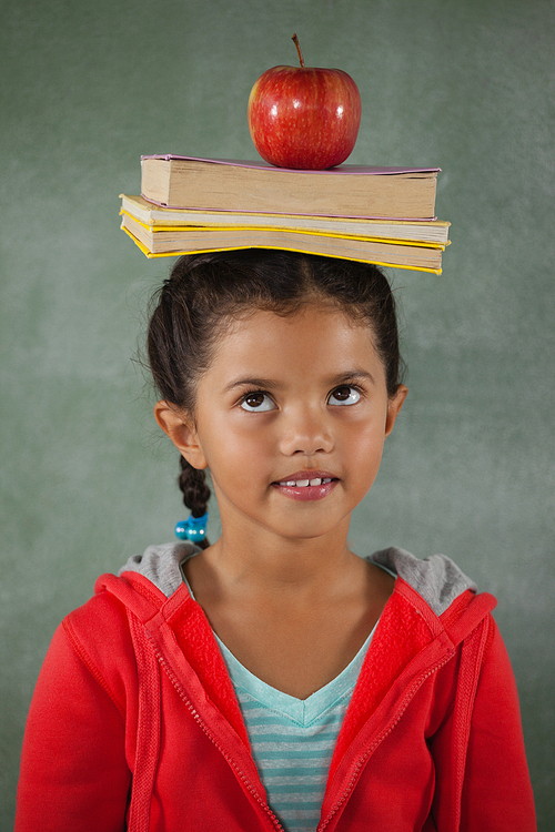 Young girl balancing books and apple on her head against chalk board