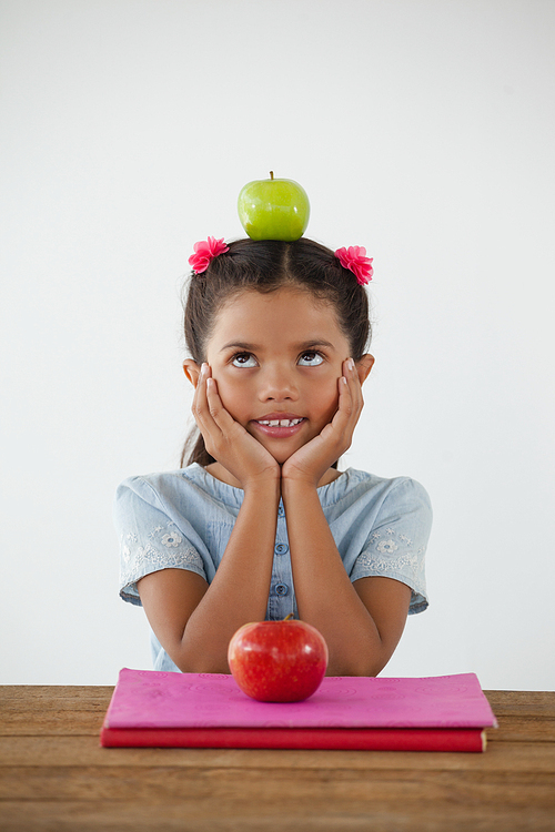 Adorable schoolgirl sitting with green apple on her head against white background