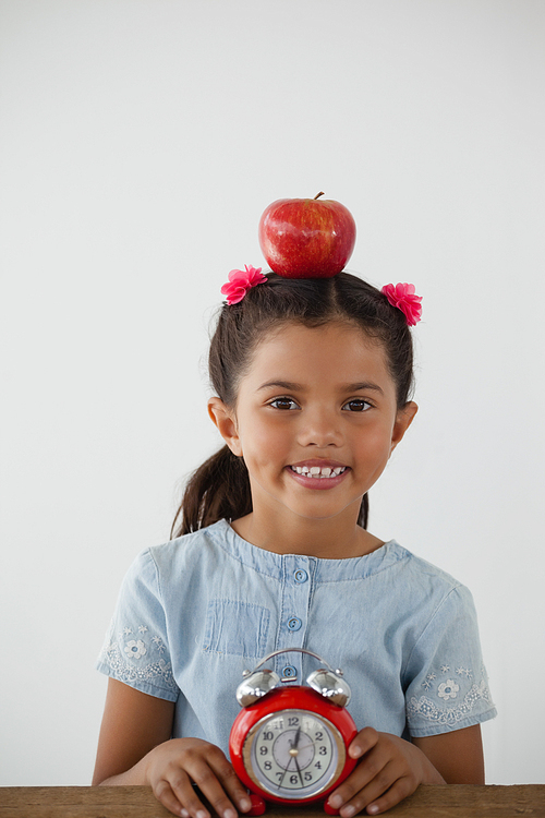 Schoolgirl sitting with apple on her head and alarm clock against white background