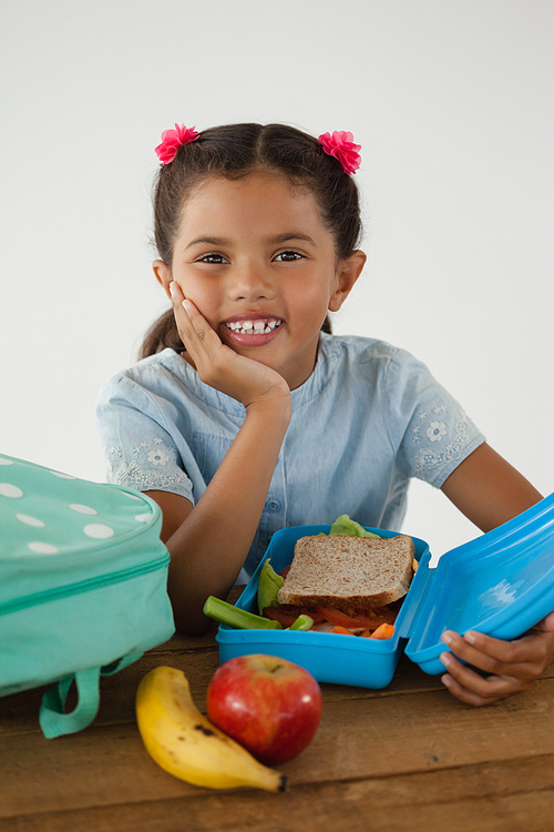 Portrait of schoolgirl sitting with tiffin box against white background