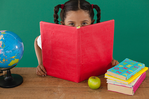 Portrait of schoolgirl hiding behind a book against green background