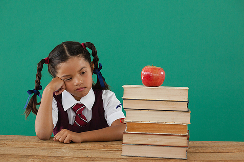 Unhappy schoolgirl looking at books stack and apple against chalkboard in classroom
