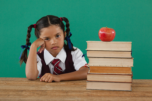 Portrait of schoolgirl sitting beside books stack with apple on top against chalkboard