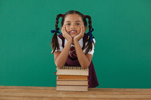 Portrait of schoolgirl leaning on books stack against chalkboard in classroom