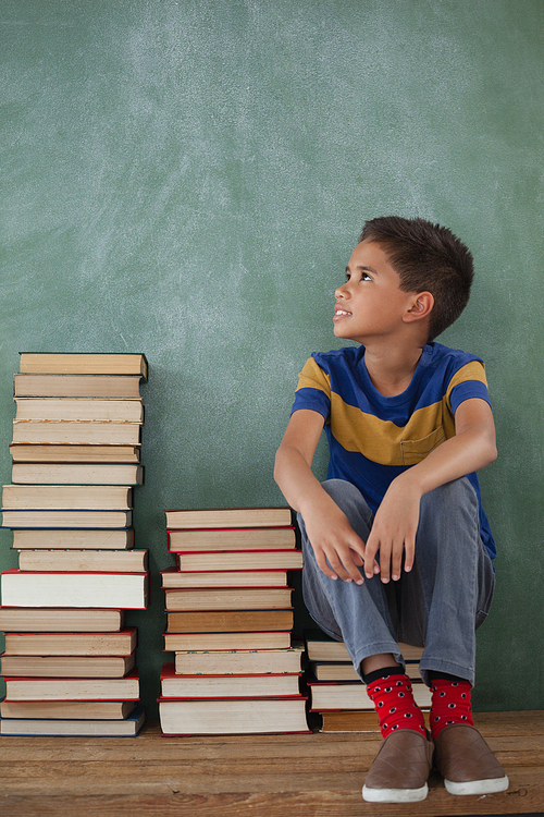 Thoughtful schoolboy sitting on books stack against chalkboard