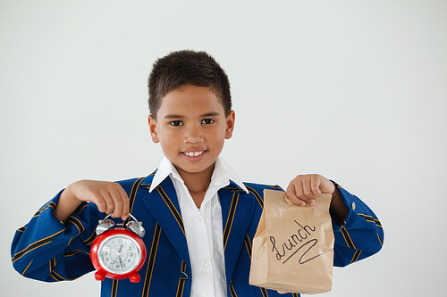 Portrait of schoolboy holding alarm clock and disposable lunch bag against white background