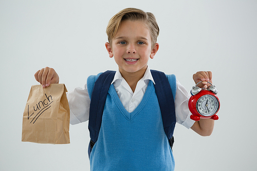 Portrait of smiling schoolboy holding alarm clock and lunch paper bag