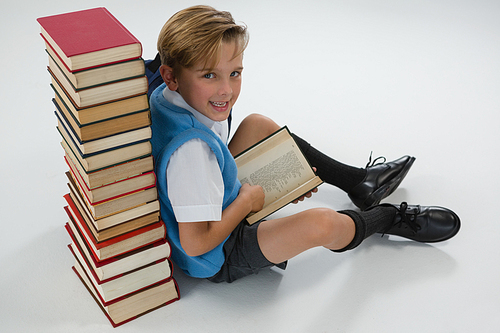 Portrait of schoolboy reading book while sitting against books stack on white background