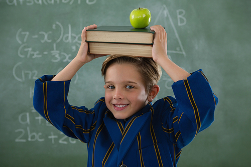 Portrait of schoolboy holding books stack with apple on head against chalkboard