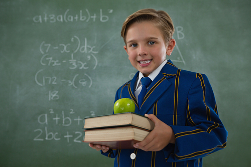 Portrait of schoolboy holding books stack with apple against chalkboard