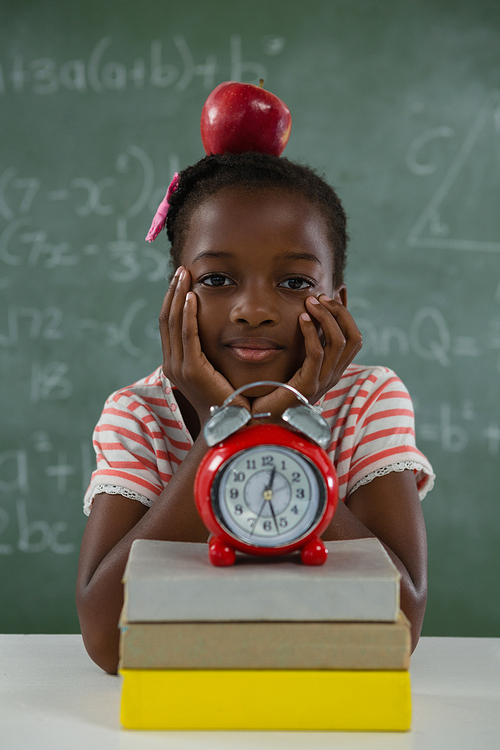 Portrait of schoolgirl sitting with red apple on her head against chalkboard