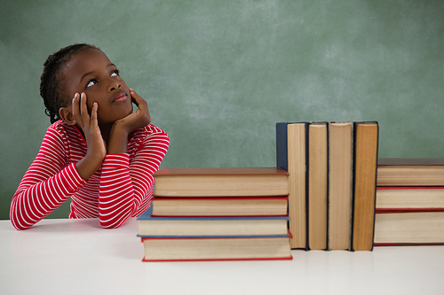Thoughtful schoolgirl sitting beside books stack against chalkboard