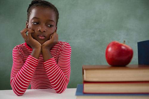 Thoughtful schoolgirl sitting beside books stack against chalkboard